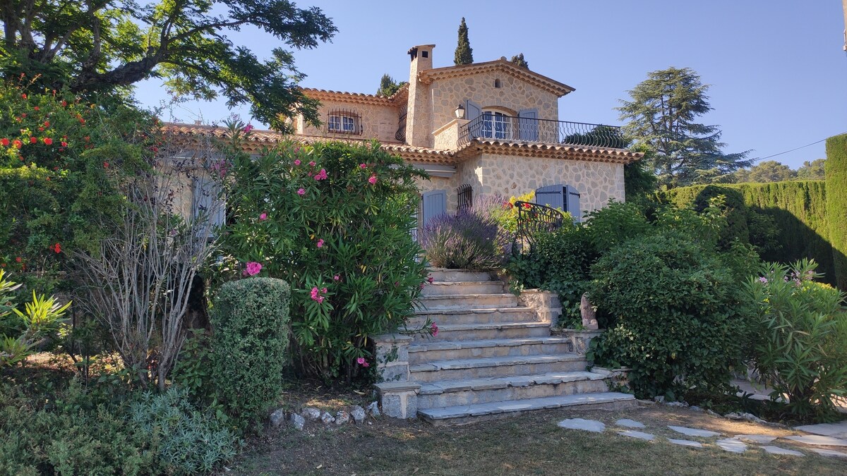 A stone villa is prominently featured, surrounded by a lush garden with vibrant flowers and greenery. Steps lead to the entrance, and trees provide shade. The structure showcases a traditional design with a terrace, offering a picturesque view of the serene landscape.