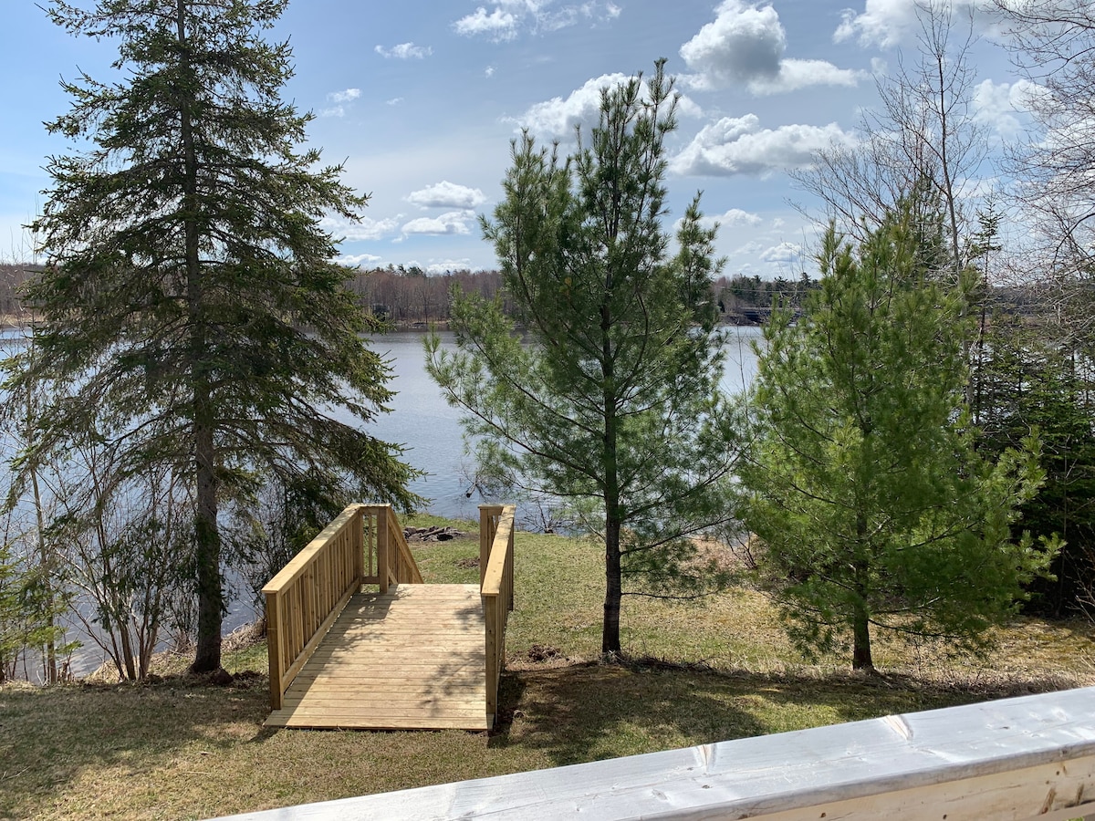 A wooden deck extends towards the tranquil waters of the West River, surrounded by lush green grass and tall pine trees. The clear sky above features fluffy white clouds, creating a serene outdoor space ideal for relaxation.