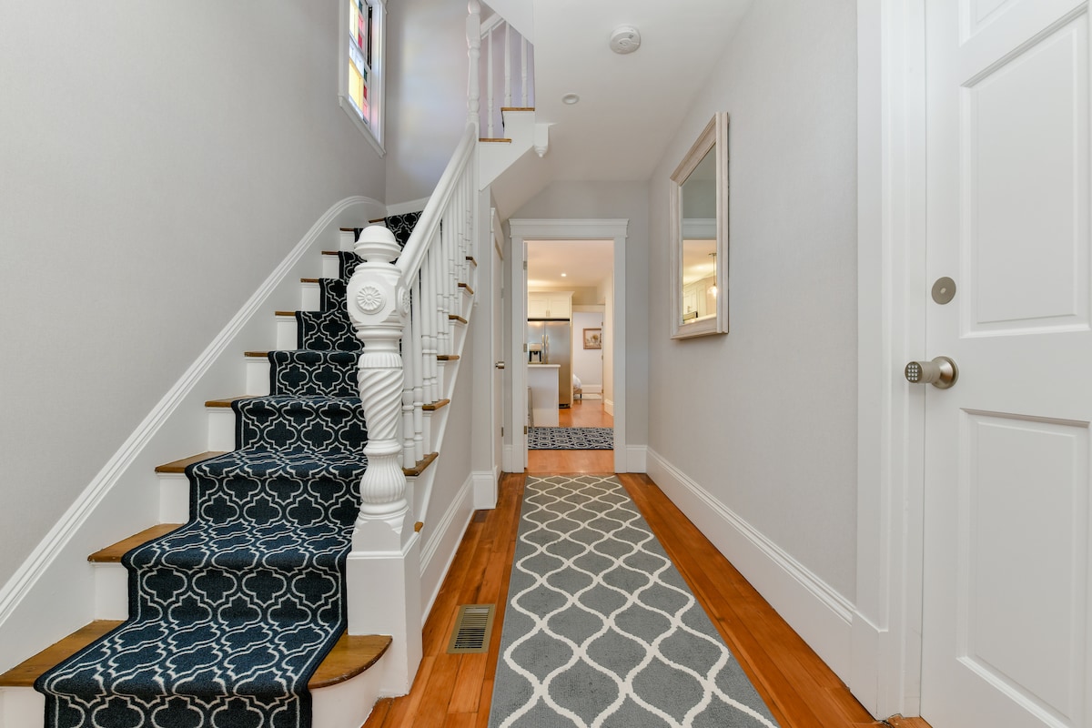 A well-lit hallway is featured, showcasing elegant stair steps adorned with a patterned navy runner. A light-colored wall and a large mirror enhance the sense of space. A patterned area rug runs along the wooden floor, leading towards an open area at the end.