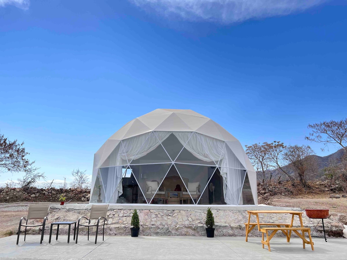 A geodesic dome structure is surrounded by a spacious outdoor area, featuring a stone patio and simple wooden furniture. Transparent panels allow for natural light, while minimal greenery and mountain views are visible in the background under a clear blue sky.