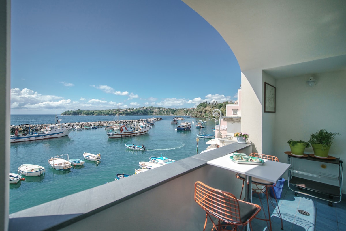 A terrace view features an arrangement of fishing boats anchored in clear blue waters, surrounded by green hills. A small table and chairs are positioned on the terrace, with potted plants adding a touch of nature. The expansive sky is partly cloudy, enhancing the serene atmosphere.