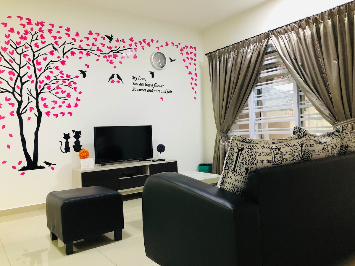 The living room features a contemporary design with an L-shaped sofa and a smart TV positioned on a low console. A decorative wall mural with pink leaves adds character, while elegant curtains frame the window, allowing natural light to create a welcoming ambiance.