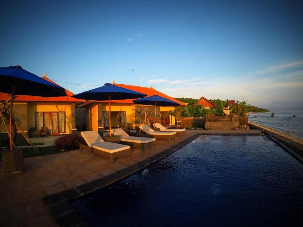 A serene swimming pool reflects the early morning sky, surrounded by lounge chairs and umbrellas. The cottage with orange rooftops sits beside the sea, enhancing the coastal setting. Exotic plants frame the area, contributing to a peaceful atmosphere by the water's edge.