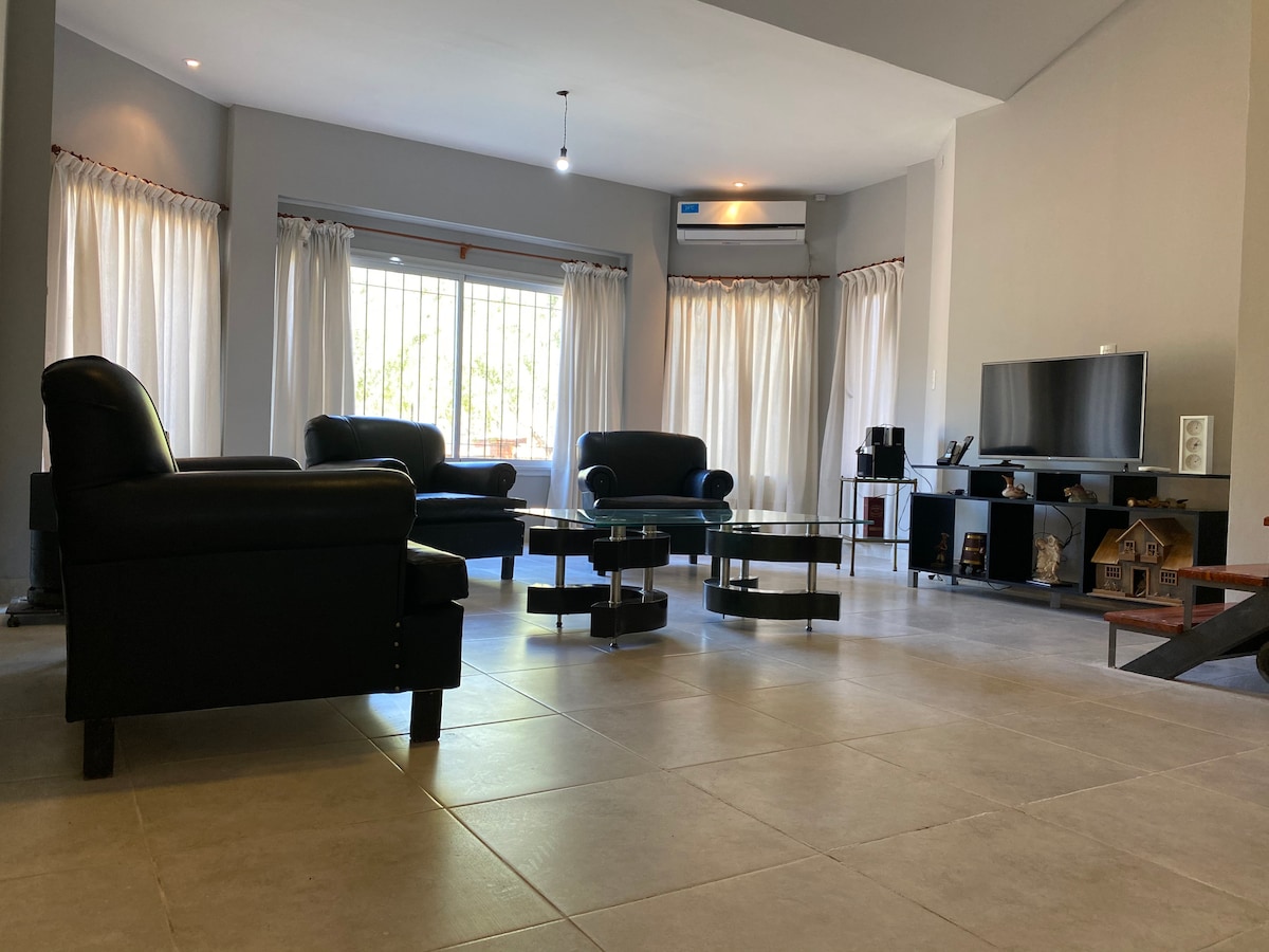 A spacious living area is characterized by modern furnishings, including black leather chairs and glass tables. Natural light filters in through large windows with white curtains, adding brightness to the neutral-toned walls. A television unit is positioned against one wall, complementing the décor.