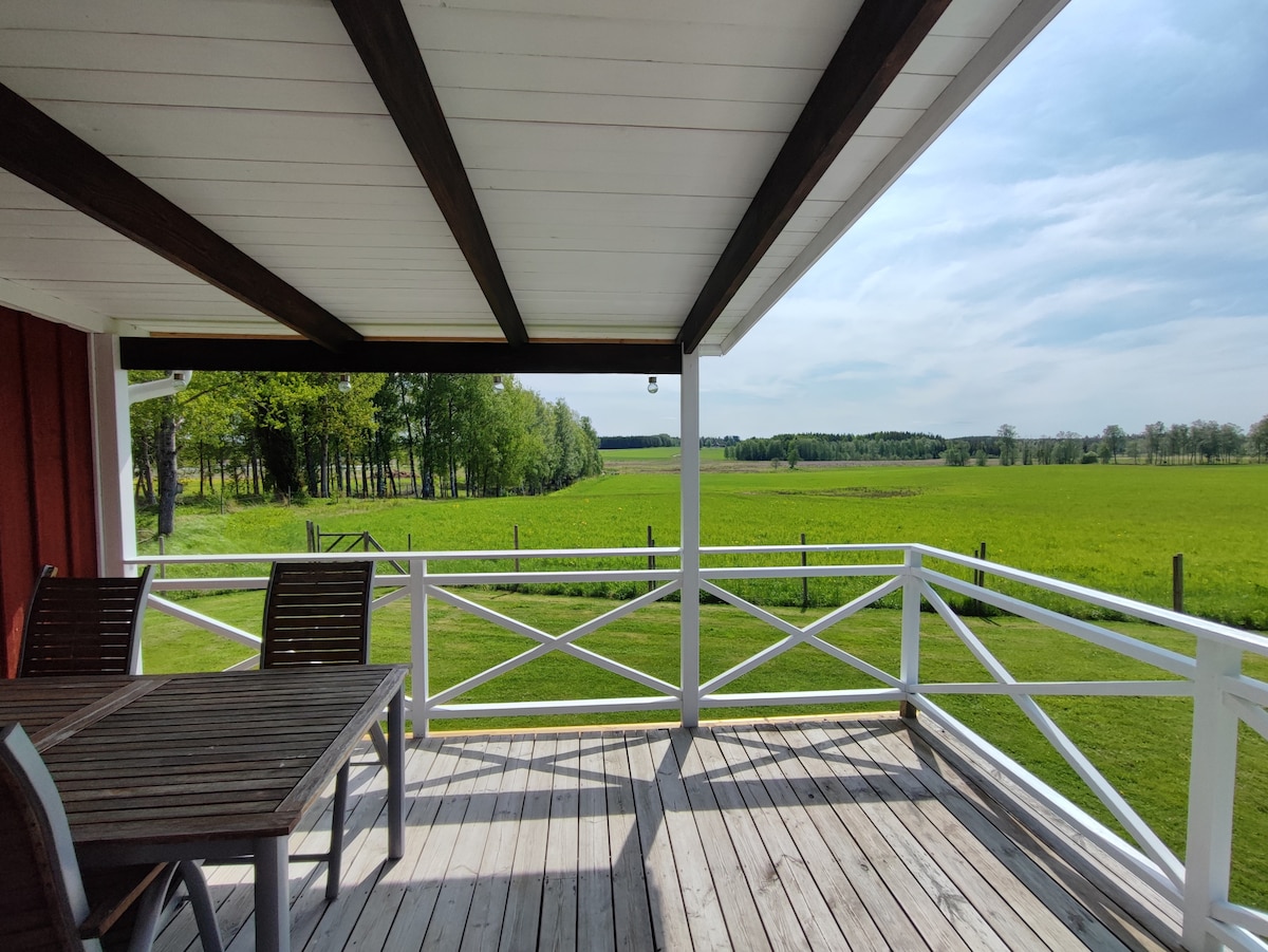 A spacious outdoor patio features a wooden table and chairs under a roof. Lush green fields stretch out beyond a white fence, framed by trees in the distance. Soft sunlight illuminates the area, creating a relaxed ambiance.