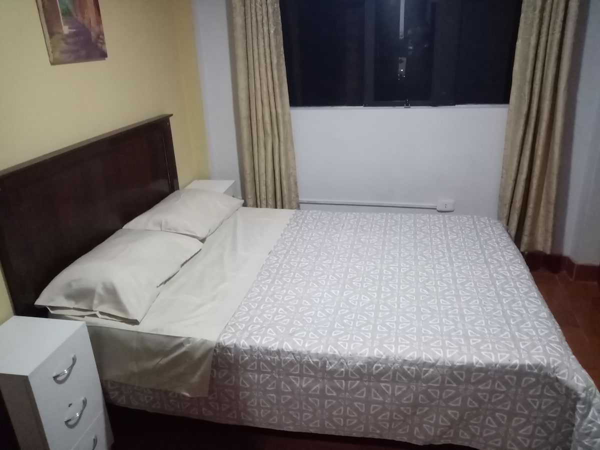 A sleeping area is presented with a spacious bed dressed in a light patterned coverlet. Soft pillows are arranged neatly on the bed, while a small nightstand is positioned beside it. Natural light enters through a large window, complemented by light-colored curtains.