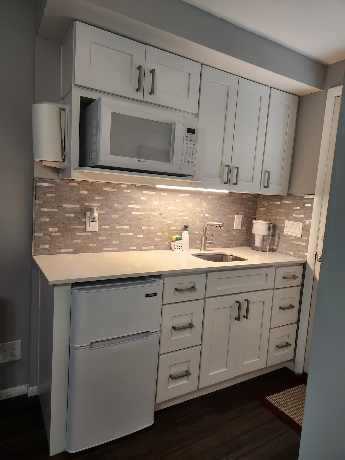 A compact kitchenette features modern white cabinetry and a light gray tiled backsplash. Essential appliances include a microwave, a small refrigerator, and a sink, complemented by a countertop for meal preparation. Soft lighting enhances the functionality and cleanliness of the space.