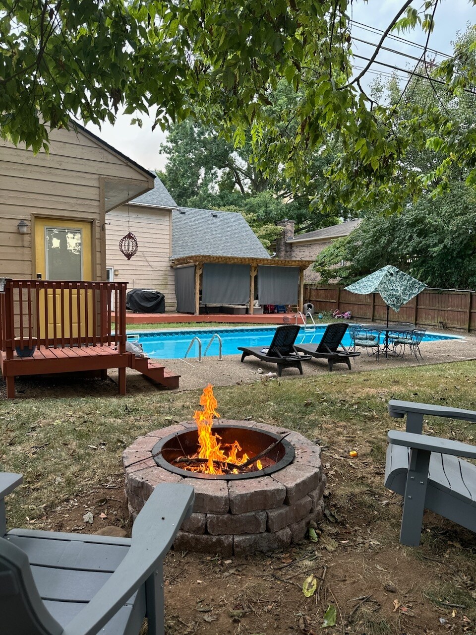 A private outdoor area features a stone firepit surrounded by gray chairs, providing a cozy setting for relaxation. In the background, a heated saltwater pool is framed by lounge chairs and a shaded seating area, amidst lush greenery and a wooden deck.