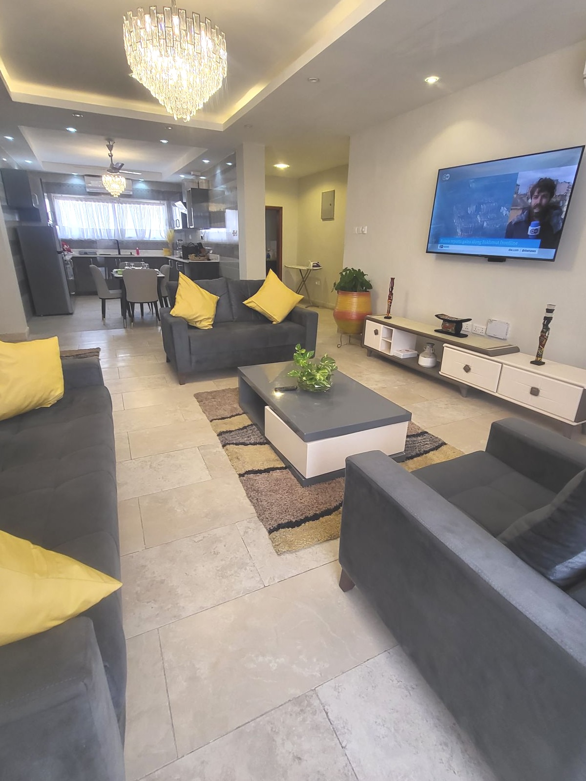 A spacious living area features grey sofas adorned with yellow cushions, paired with a modern coffee table. A flat-screen television is mounted on the wall, while natural light brightens the space through large windows. A dining area is visible in the background.