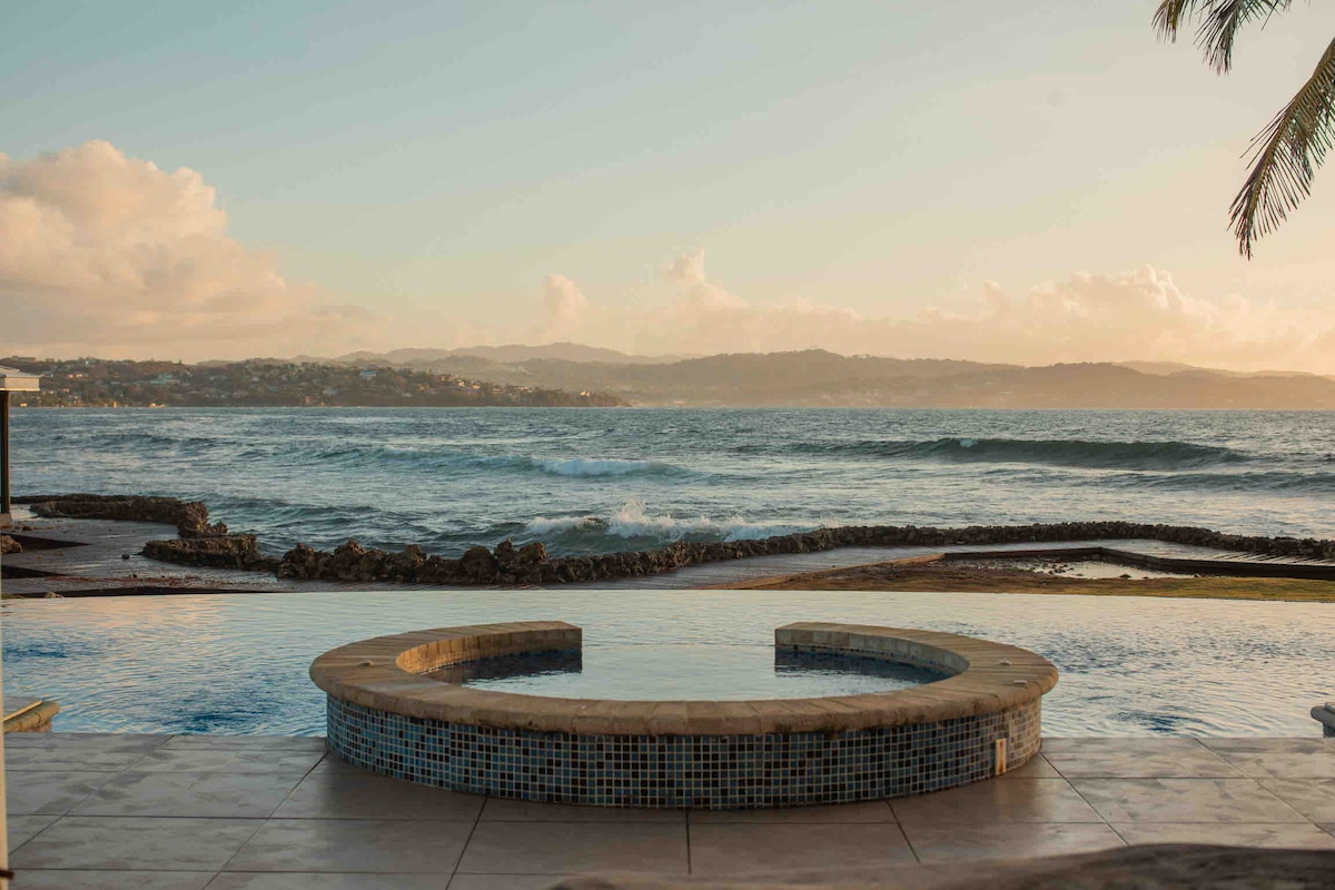 An infinity pool merges with the ocean, featuring a round heated jacuzzi at the foreground. Waves gently lap against the rocky shoreline, while distant hills are visible under a warm sunset sky.
