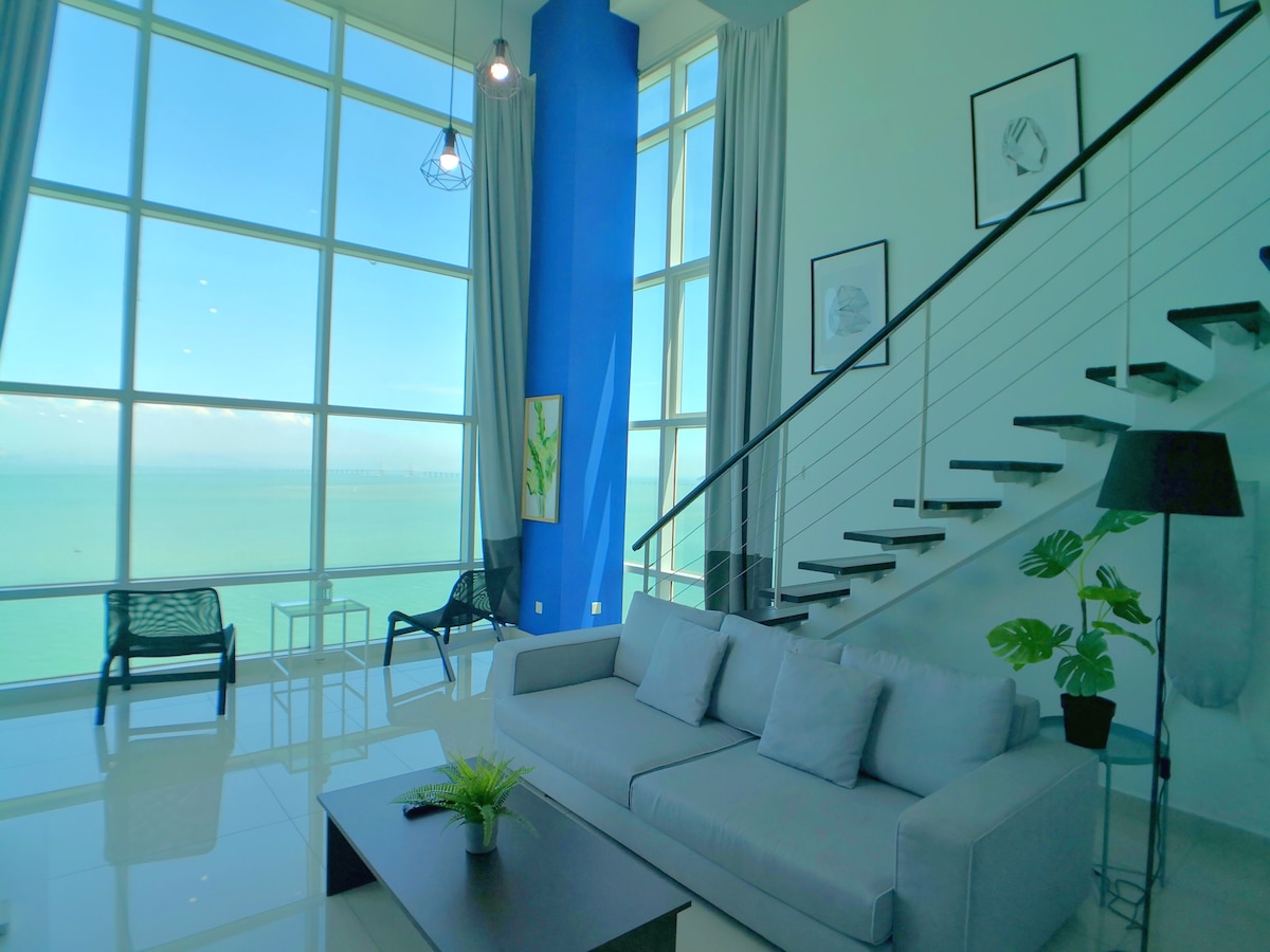 A spacious living area features large floor-to-ceiling windows that offer expansive sea views. The room includes a light-colored sofa and a small coffee table. A staircase extends upward, complemented by minimalist artwork on the wall and modern decor elements.