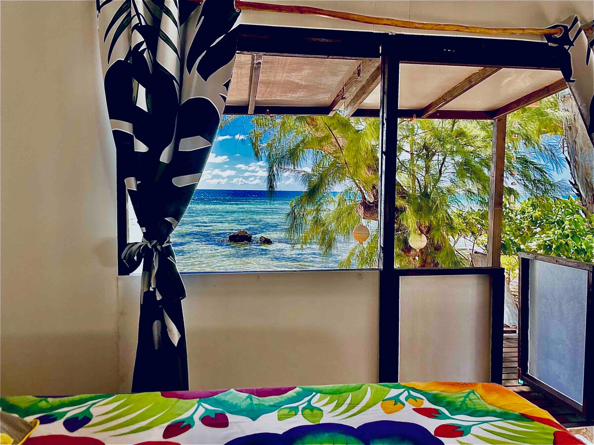 The image captures a bright interior view from a bungalow, revealing a stunning seascape through large windows. Colorful tropical foliage is framed against the backdrop of the ocean, with gentle waves and distant rocks visible. A patterned bedspread adds a splash of color to the scene.