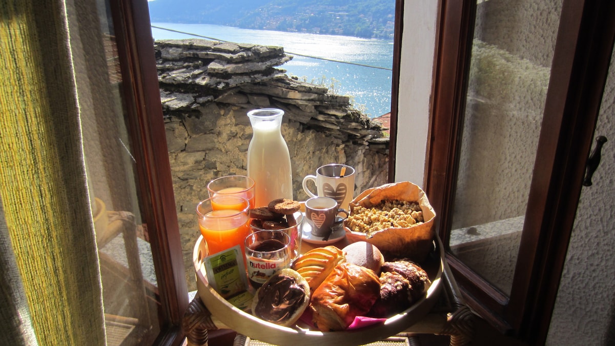 A breakfast tray is positioned on a window sill, showcasing a variety of foods including pastries, granola, and cups of coffee and juice. The picturesque lake view through the window enhances the inviting scene.
