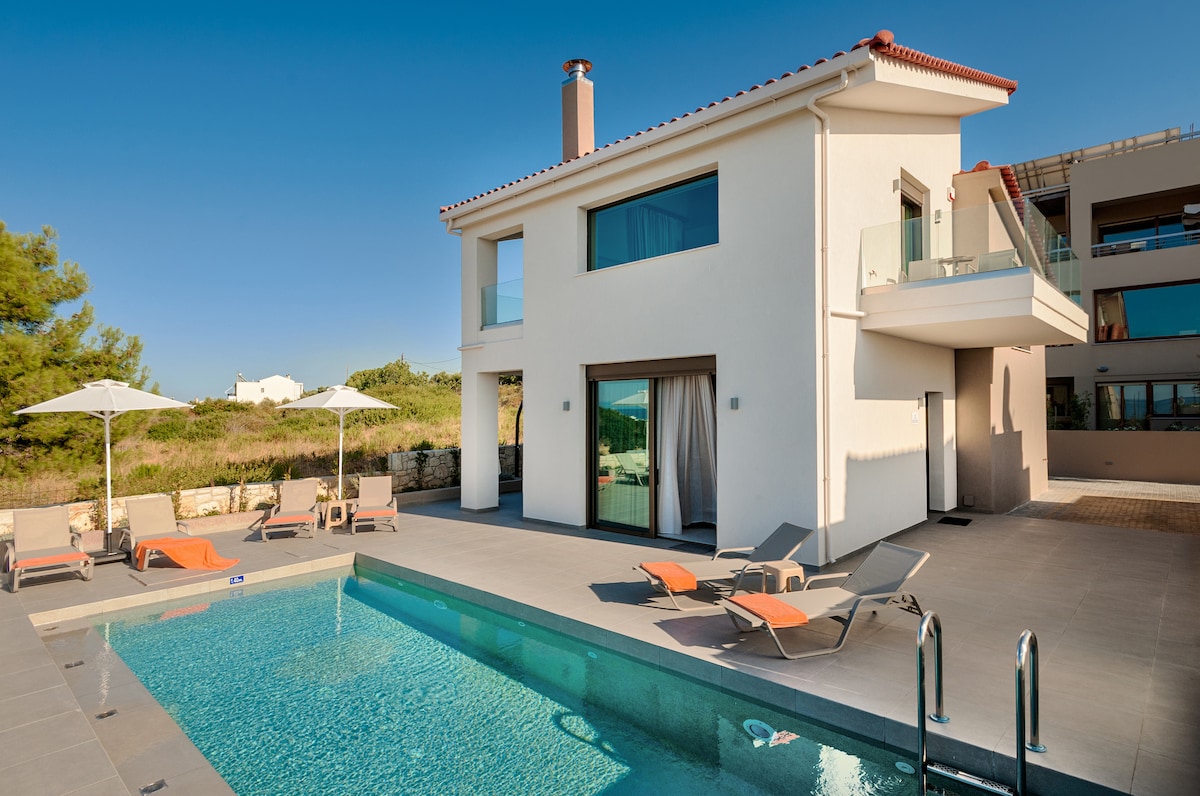 A luxury villa features a modern exterior with a spacious layout. A private swimming pool is showcased, bordered by sunbeds and umbrellas, inviting relaxation. Large windows provide ample natural light, complimenting the clean lines and contemporary design of the villa.