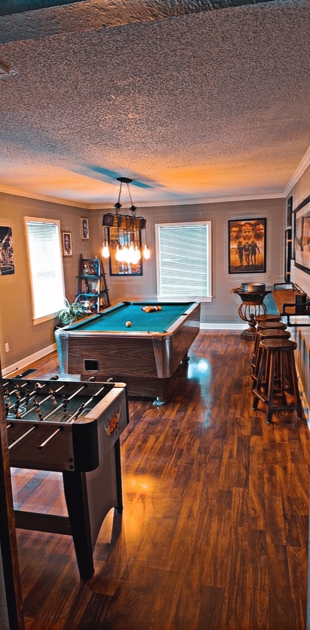 The game room features a pool table centered in the space, complemented by a foosball table and several bar stools. Natural light from windows illuminates the warm wooden flooring, enhancing the inviting atmosphere of the area designed for entertainment.