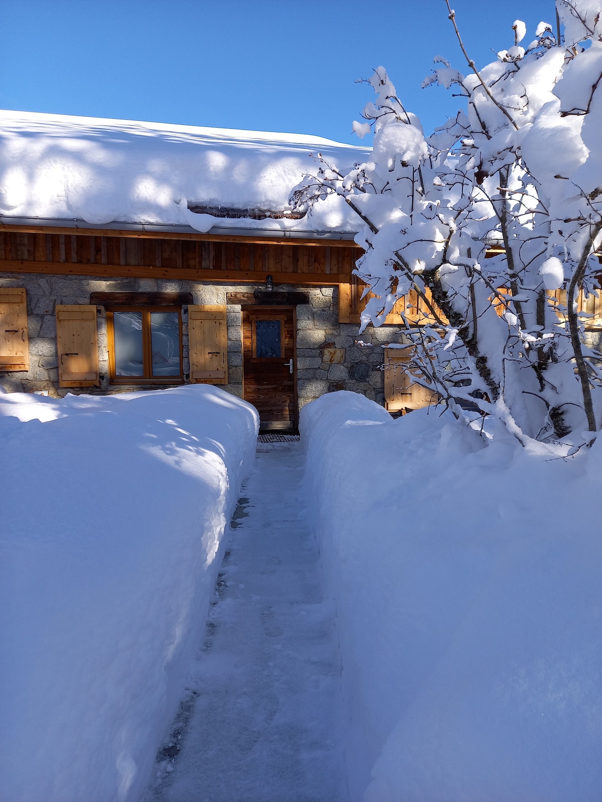 A pathway bordered by snowdrifts leads to the entrance of a rustic mountain farmhouse. Sunlight reflects off the snow, illuminating wooden shutters and a sturdy door. Snow-covered trees frame the scene, creating a serene and wintry atmosphere.