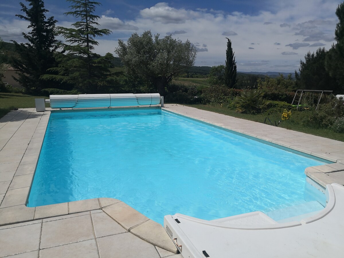 An outdoor swimming pool is featured in a private area, framed by landscaped gardens and lush greenery. The clear blue water reflects sunlight, while a paved patio surrounds the pool, providing space for relaxation. Scenic views of the countryside are visible in the background.
