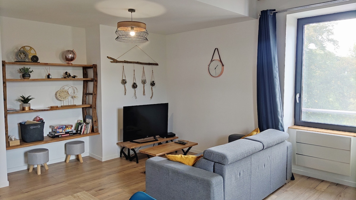 A comfortable living area features a grey sofa facing a wooden coffee table and TV. Decorative shelves display plants and books. Two small stools are situated nearby. A large window allows soft natural light to fill the space, with light curtains framing the view.
