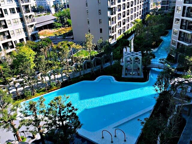 The image features a large outdoor swimming pool surrounded by lush greenery and modern condominium buildings. Sunlight reflects on the water's surface, creating a tranquil atmosphere. Lounge chairs are positioned along the poolside, providing ample space for relaxation.
