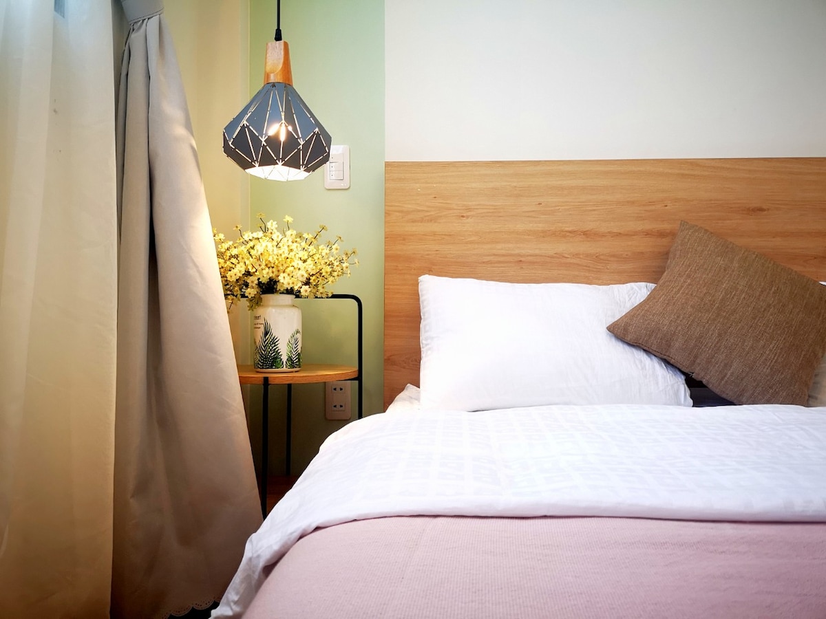 A serene bedroom corner features a soft mattress on a large bed, complemented by high-quality bedding. A bedside table holds a decorative vase with flowers. The space is illuminated by a modern pendant light, casting a warm glow over the light-colored walls.
