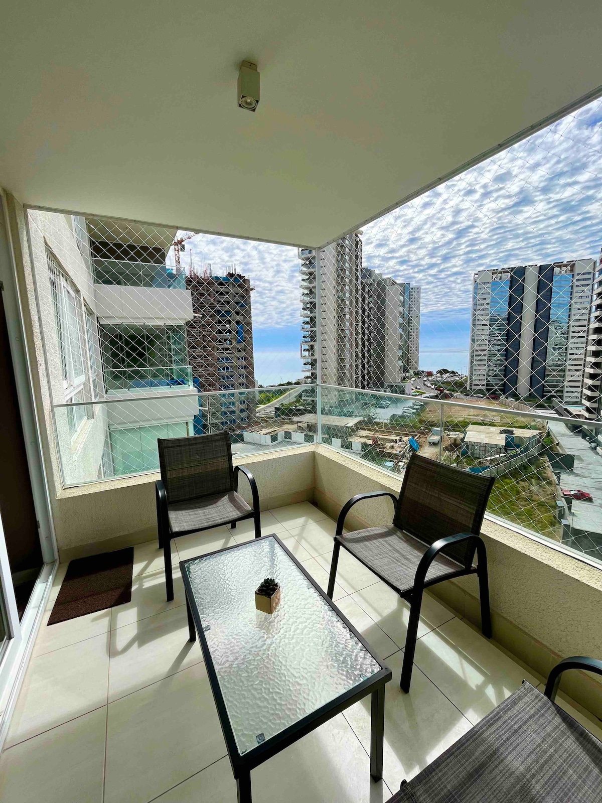 A balcony area is shown, featuring a glass table surrounded by four chairs. The view includes city buildings and a glimpse of the ocean, with the sky filled with scattered clouds. The space is enclosed with a safety netting for security.