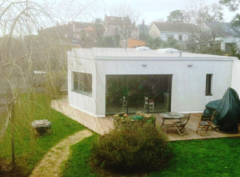 A modern, single-story home is set within a spacious garden. Large glass windows provide views into the interior, while outdoor seating is arranged on a wooden terrace. Lush green grass and a winding path enhance the outdoor space, contributing to a tranquil setting.