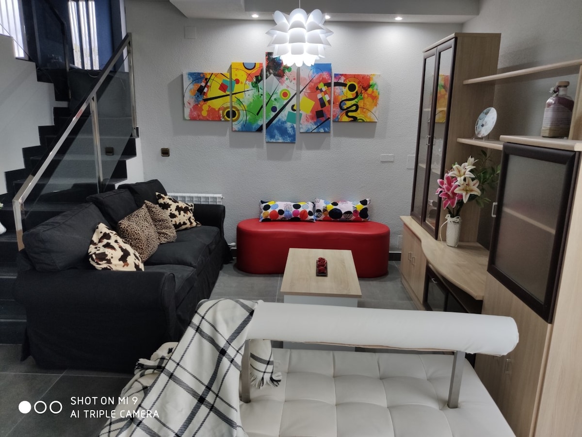 A spacious living room displays a mix of seating, including a dark sectional couch and a vibrant red sofa. Colorful artwork adorns the wall above a wooden coffee table, while a decorative blanket rests on the arm of a sleek chair. Natural light fills the area through a nearby window.