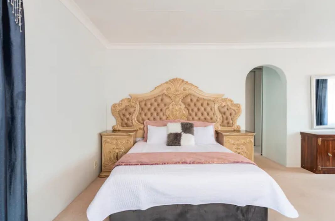 A spacious sleeping area features an ornate headboard crafted from a soft, tufted material. The bed is adorned with decorative pillows and a light bedspread. Neutral walls create a serene atmosphere, while soft carpet covers the floor.