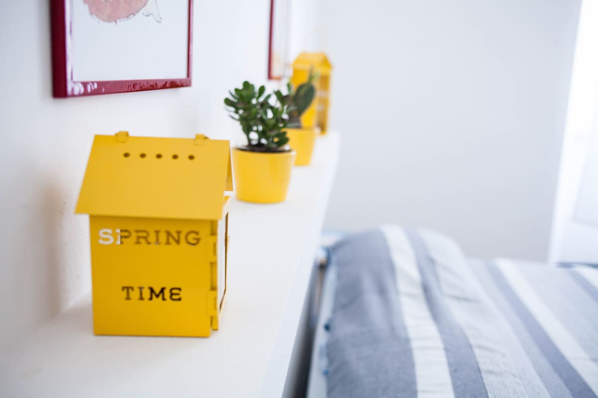 A bright shelf displays a small yellow house decor labeled 'SPRING TIME' next to a potted succulent. The background features a striped bedding, providing a lively touch to the clean and well-lit space.