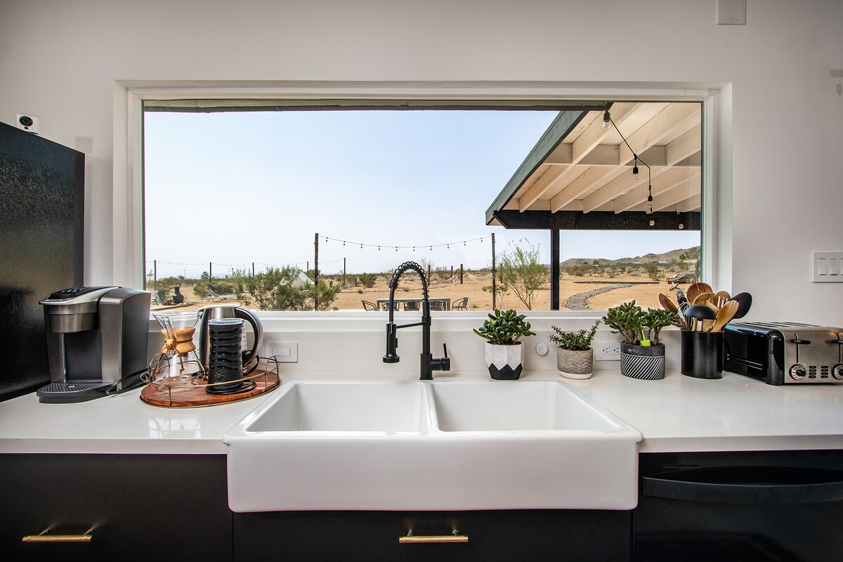 A modern kitchen is displayed with a large farmhouse sink positioned beneath an expansive window, providing a view of the surrounding desert landscape. The counter features kitchen appliances, utensils, and potted plants, creating a functional cooking space with natural light.