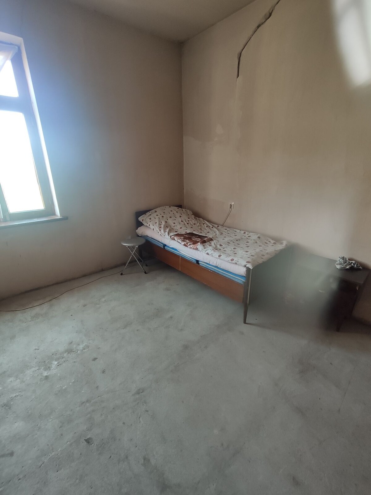 A simple room is presented with bare walls and a concrete floor. A single bed with a patterned blanket is positioned against one wall, accompanied by a small side table nearby. A window allows natural light to enter the space.
