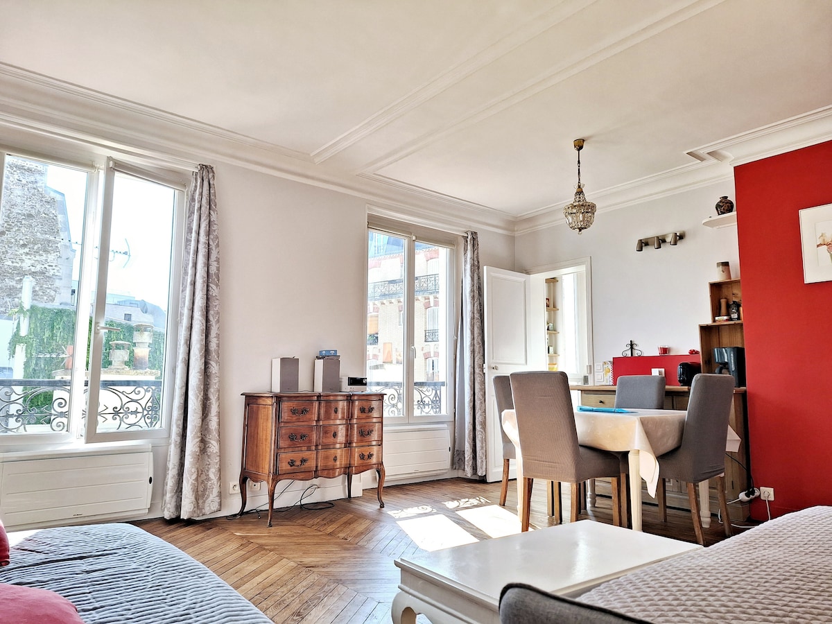 A spacious living area features large windows allowing natural light to enter. An elegant wooden dining table surrounded by gray chairs is situated adjacent to a rich red wall. Vintage furniture and decorative elements contribute to the room's charm, reflecting a blend of comfort and style.