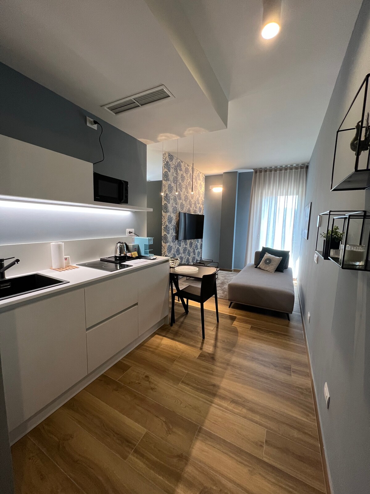The image reveals a contemporary kitchenette equipped with sleek white cabinetry and essential appliances, including a coffee maker and microwave. A small dining table with two chairs is positioned nearby, and a cozy seating area features a convertible sofa, with a window allowing natural light to fill the space.