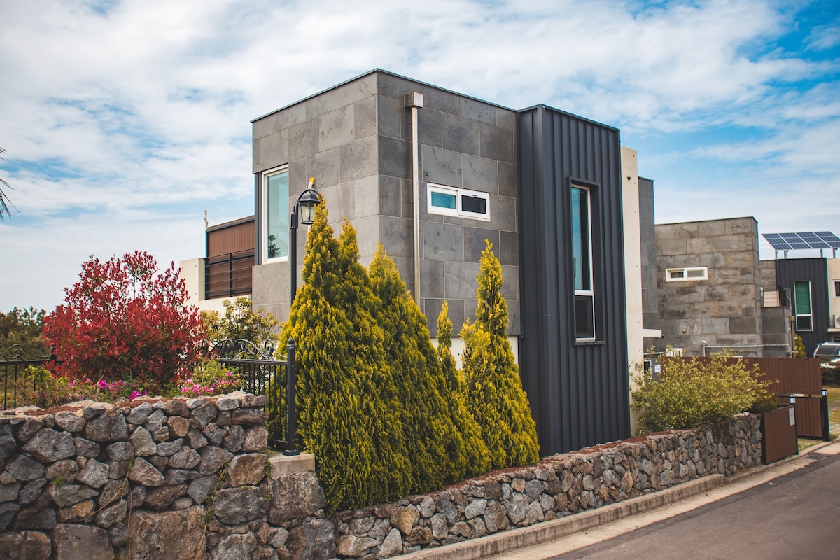 A modern two-story house with a blend of stone and metal exteriors is set against a backdrop of greenery. Neatly trimmed conifer trees flank the property, while colorful flowering shrubs are visible near the stone wall enclosing the yard.