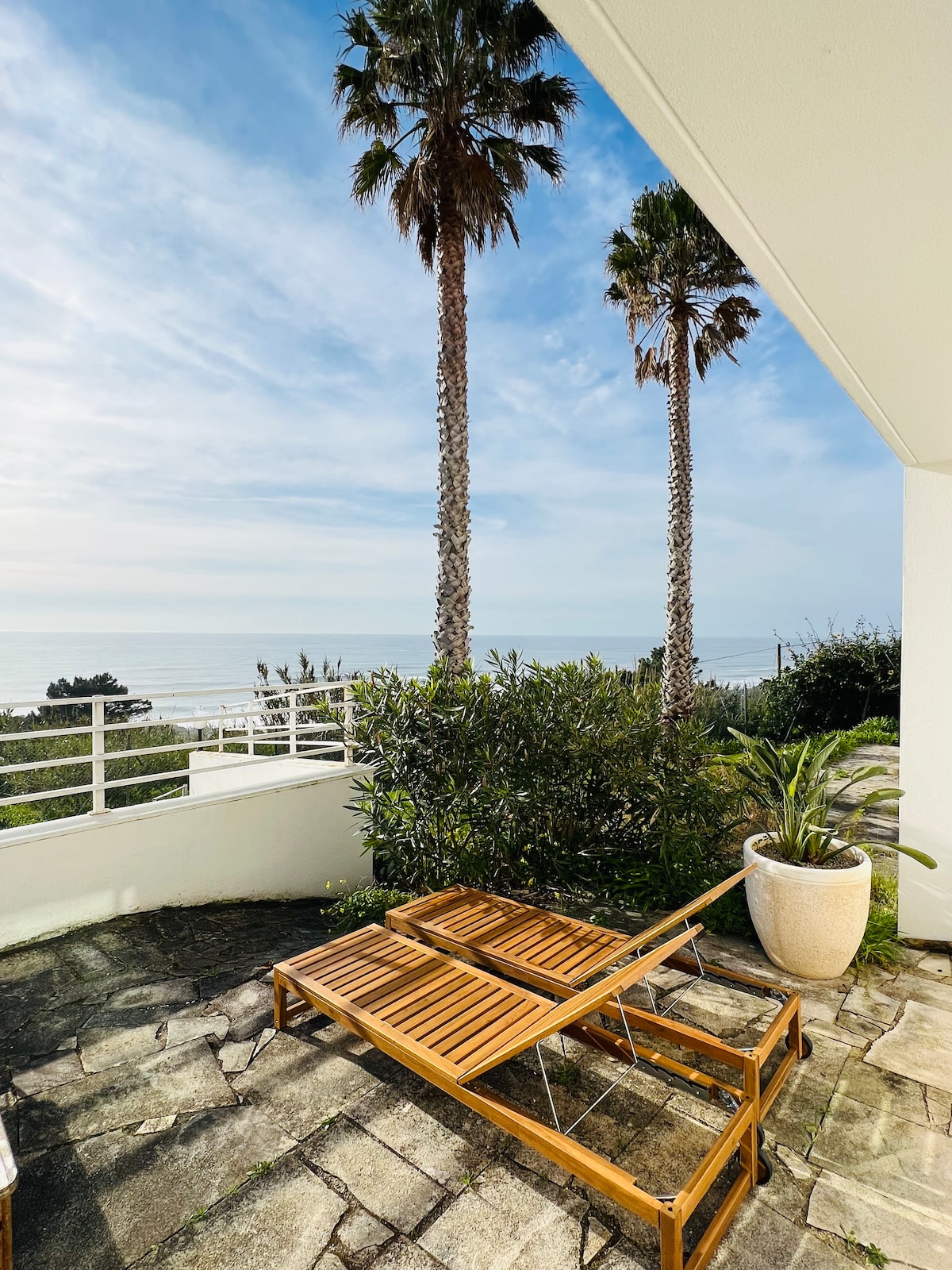 A private terrace features two wooden lounge chairs positioned on a stone patio, overlooking a serene view of the ocean. Tall palm trees and lush greenery create a relaxed outdoor ambiance, with a clear sky visible in the background.
