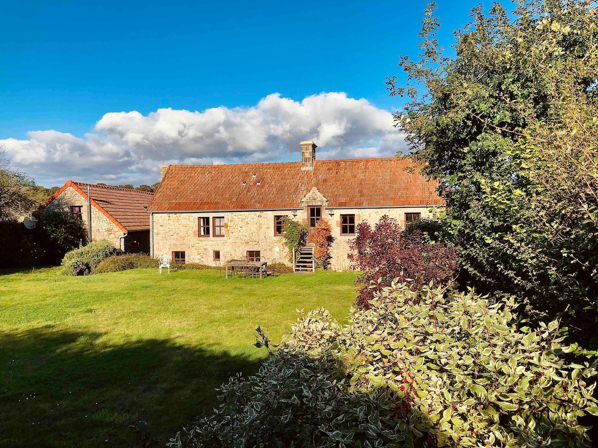 The charming cottage is set in a spacious garden, surrounded by greenery and mature shrubs. A stone façade is complemented by a warm-colored roof and multiple windows. The entrance is framed by a small staircase leading into the inviting interior.