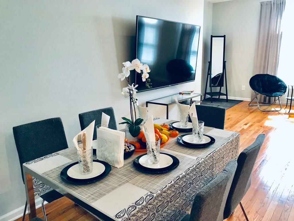 A dining area features a rectangular table set for four, adorned with a decorative table runner and plates. A large flat-screen television mounted on the wall is complemented by an accent mirror nearby. The space is filled with natural light from a window with sheer curtains.
