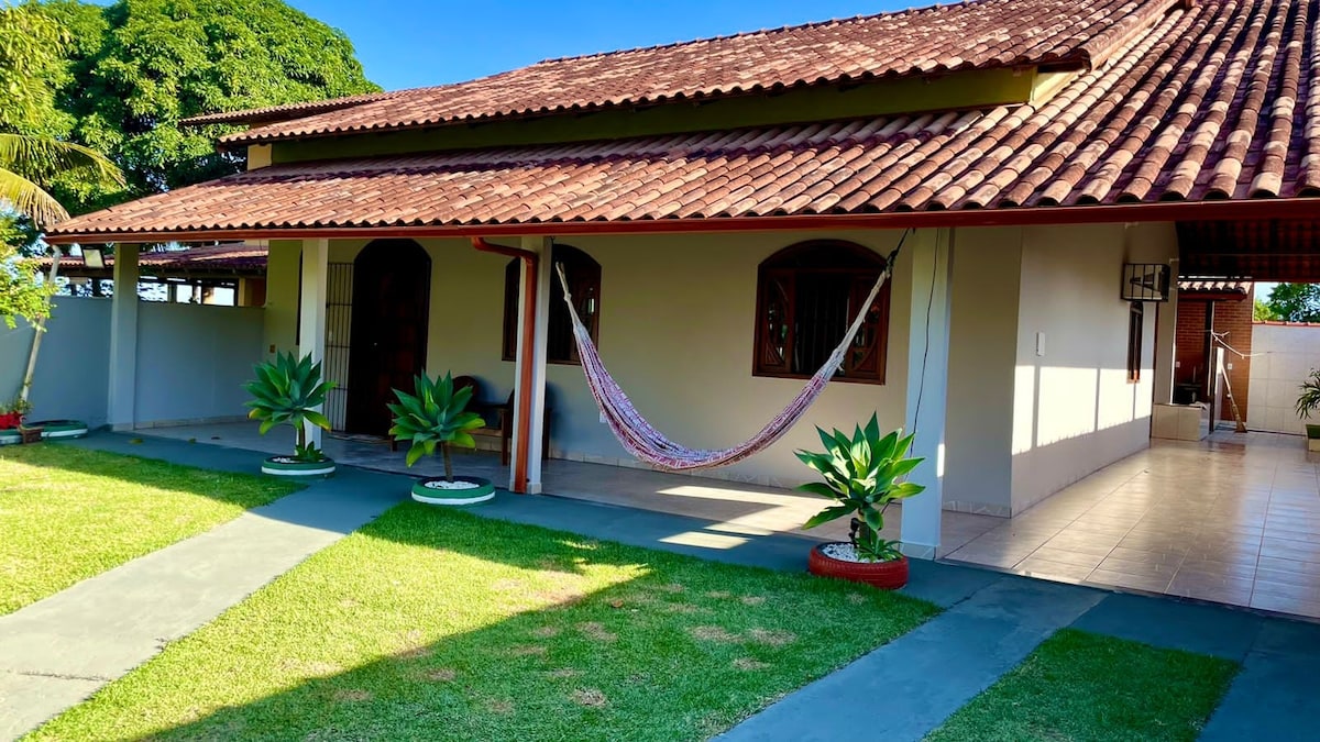 The exterior of the house is showcased with a tiled roof and large windows. A vibrant hammock is hung between two posts, complemented by potted plants. A smooth pathway leads to the entrance, surrounded by a well-maintained lawn.