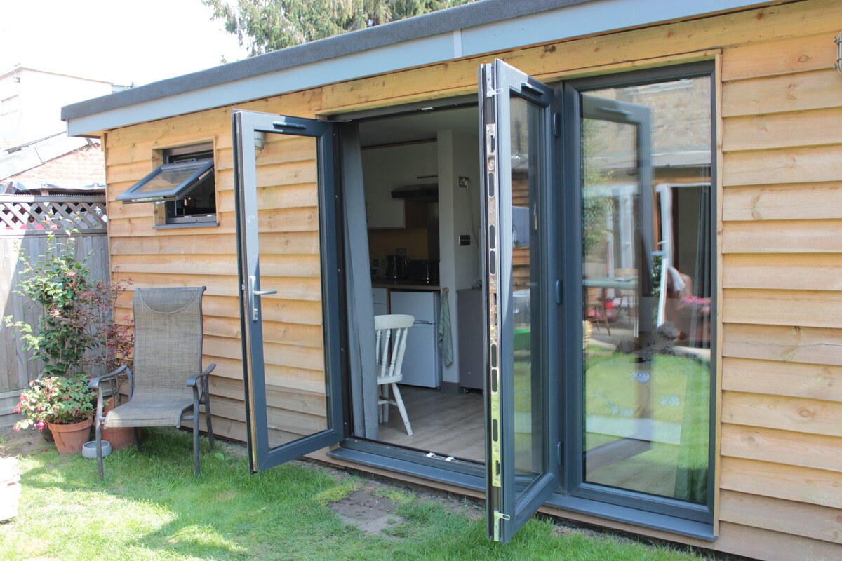 The image showcases a modern log cabin with an open door revealing a glimpse of the stylish interior. The exterior features wooden paneling, and large glass doors provide an inviting connection to the garden. A comfortable chair sits on the lawn, adding to the outdoor space.
