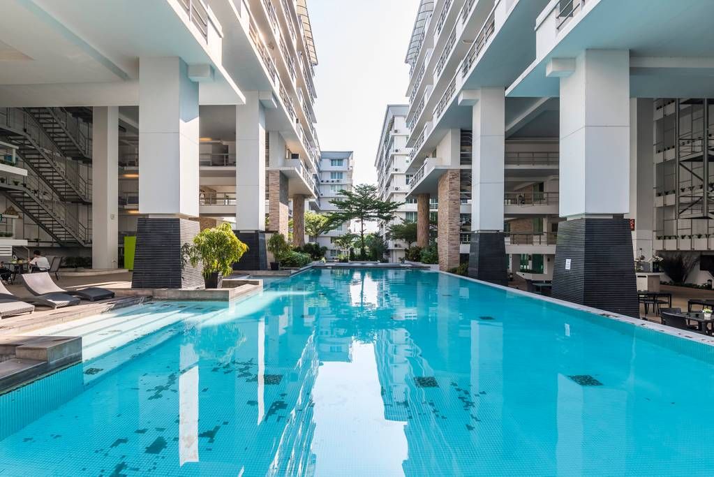 A spacious swimming pool is centered between modern buildings, surrounded by lush greenery. Sun loungers line the poolside, inviting relaxation. The clear water reflects the structure above, enhancing the sense of openness. Natural light fills the area, highlighting the contemporary design of the surroundings.