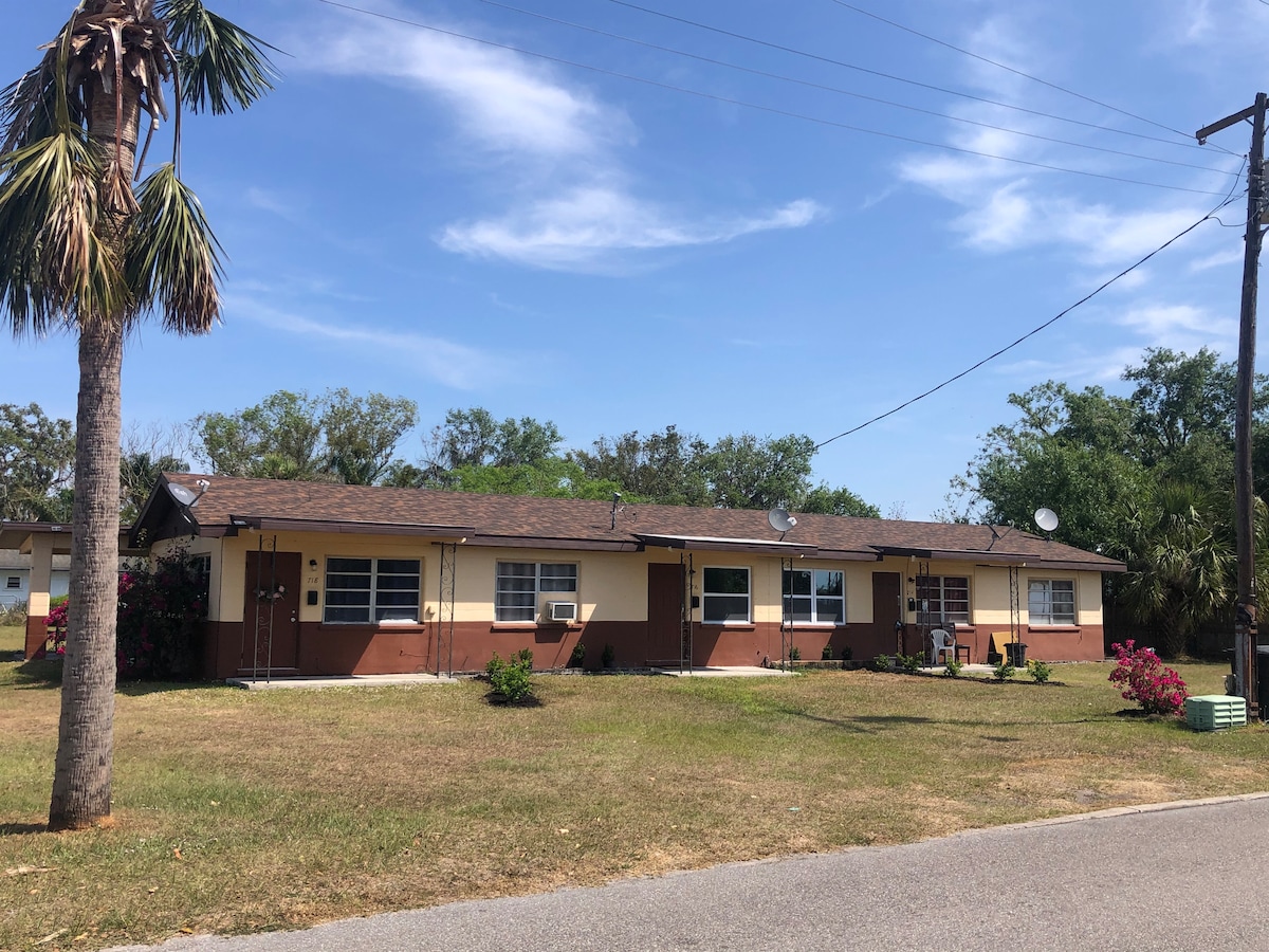 A triplex building is displayed with a light brown and beige exterior. Each unit features a small porch. Surrounding the property are green lawns and shrubs, with a tall palm tree on the left. The sky is partly cloudy, providing a bright atmosphere.