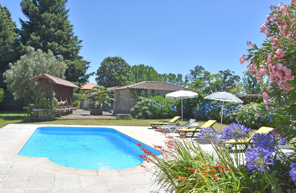 A refreshing swimming pool is surrounded by lush gardens and colorful flowers. Lounge chairs are placed around the pool, offering a relaxing space. In the background, a covered wooden area is visible, along with a building nestled among the greenery.