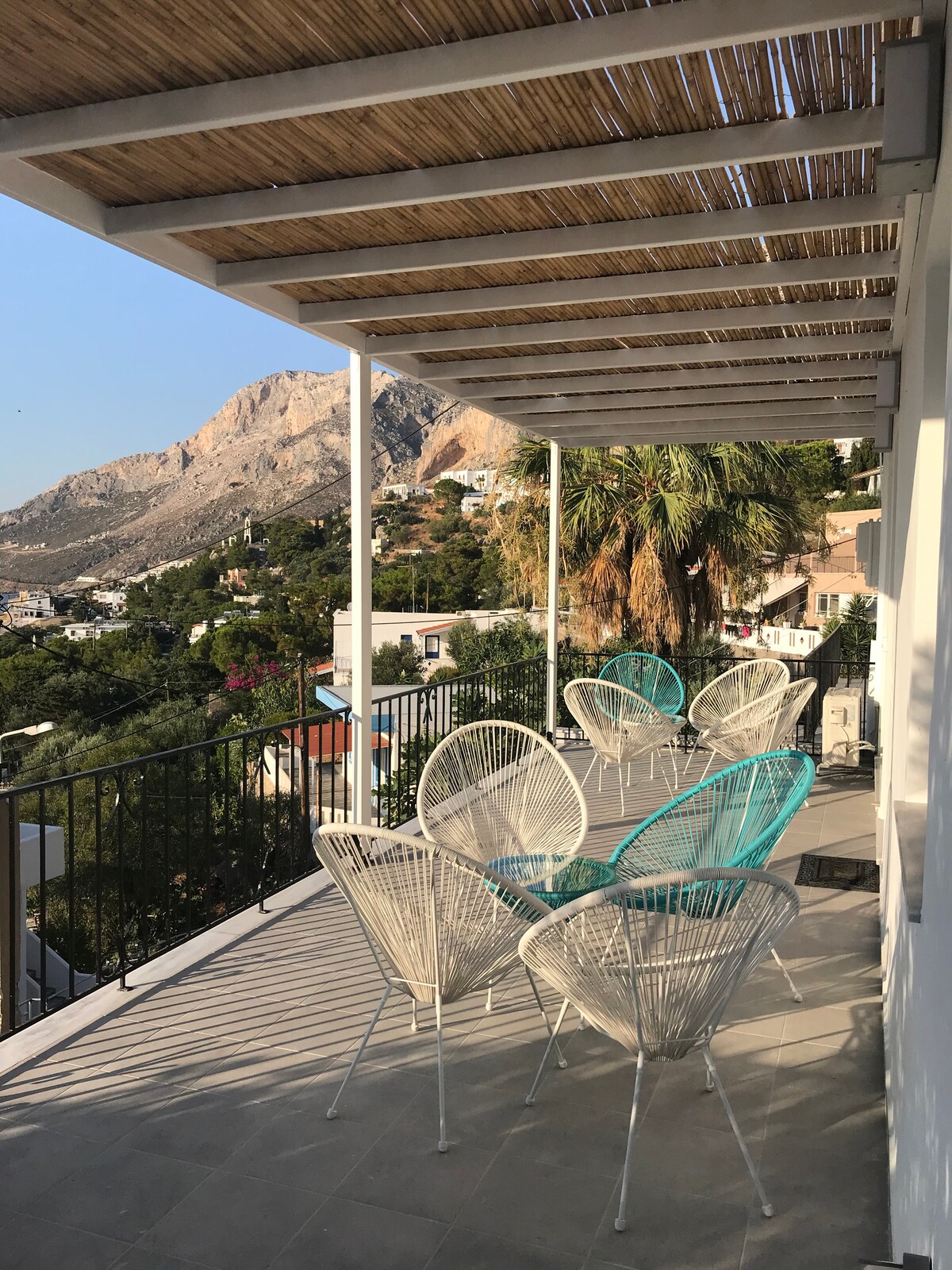 A spacious veranda featuring several white and teal outdoor chairs, offering a serene space for relaxation. The covered area provides shade while overlooking the natural landscape, characterized by hills and lush greenery, creating an inviting atmosphere for enjoying views.