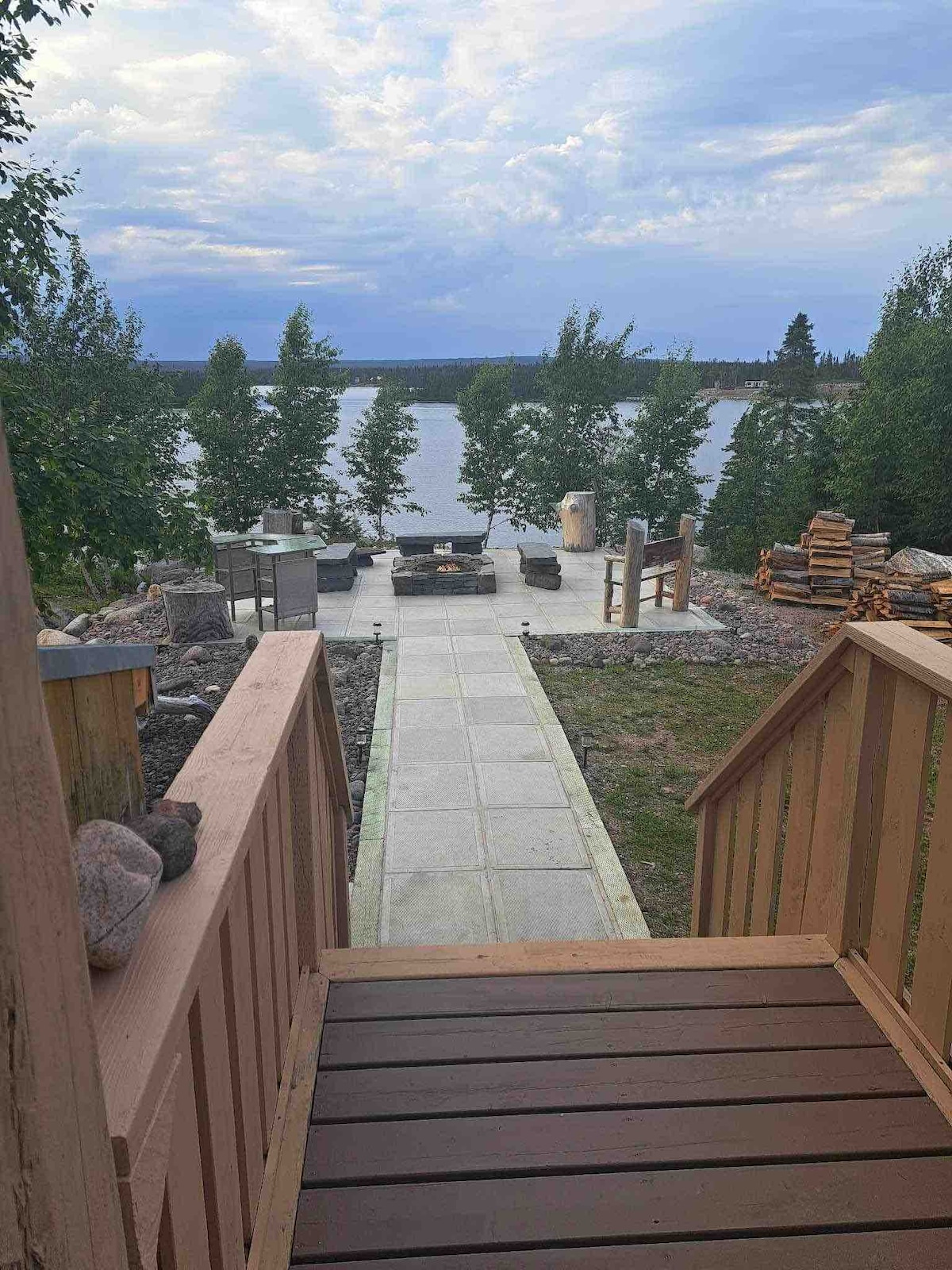 A pathway leads from the deck toward a fire pit area, framed by trees on either side. Outdoor seating arrangements are positioned around a fire pit, while a stack of firewood is visible nearby. The serene lake can be seen in the distance under a cloudy sky.
