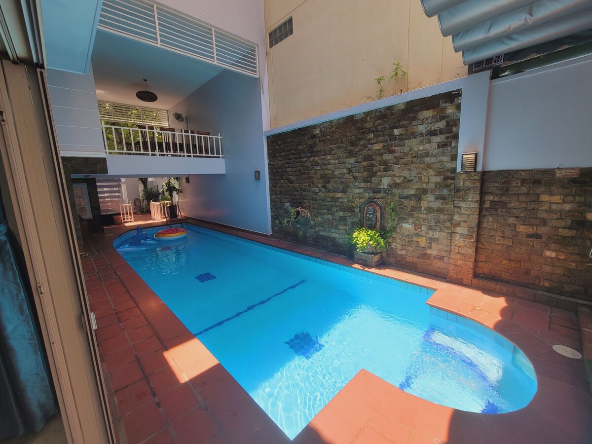 A swimming pool is highlighted in the image, featuring clear blue water and a colorful floating device. Sunlight reflects off the surface, creating a vibrant atmosphere. The surrounding walls are made of textured stone, and decorative plants are placed along the edge.