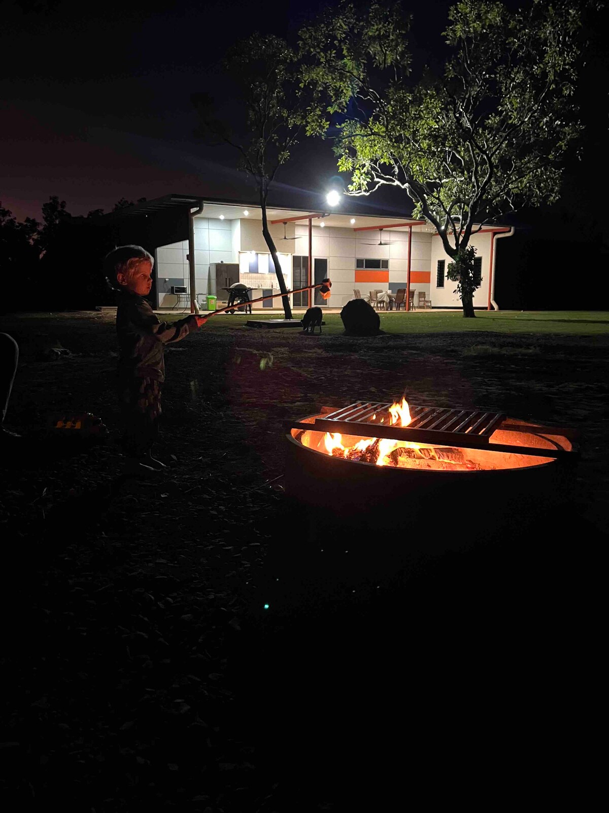 A fire pit is illuminated at night, surrounded by a grassy yard. A glowing grill sits above the flames, while the background reveals a well-lit modern structure with large windows and outdoor seating, providing a relaxed outdoor ambiance.