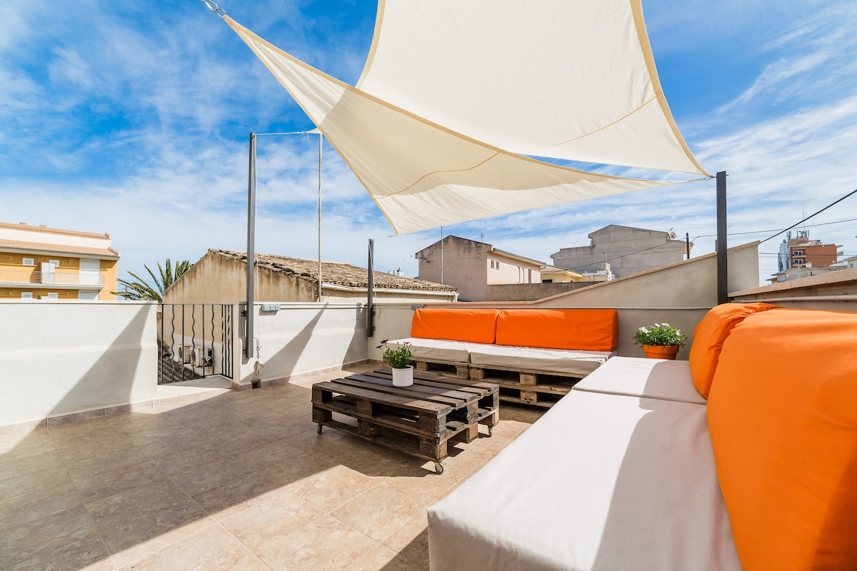 A spacious rooftop terrace is equipped with comfortable seating in orange and neutral tones. A sunshade is positioned overhead, providing a relaxed atmosphere. Potted plants add a touch of greenery, while the surrounding views are visible against a clear blue sky.