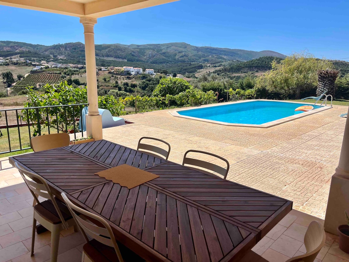 An outdoor terrace is showcased with a large wooden dining table, surrounded by several white chairs. The swimming pool is visible in the background against a scenic view of the valley and rolling hills, providing a relaxing backdrop for outdoor meals.
