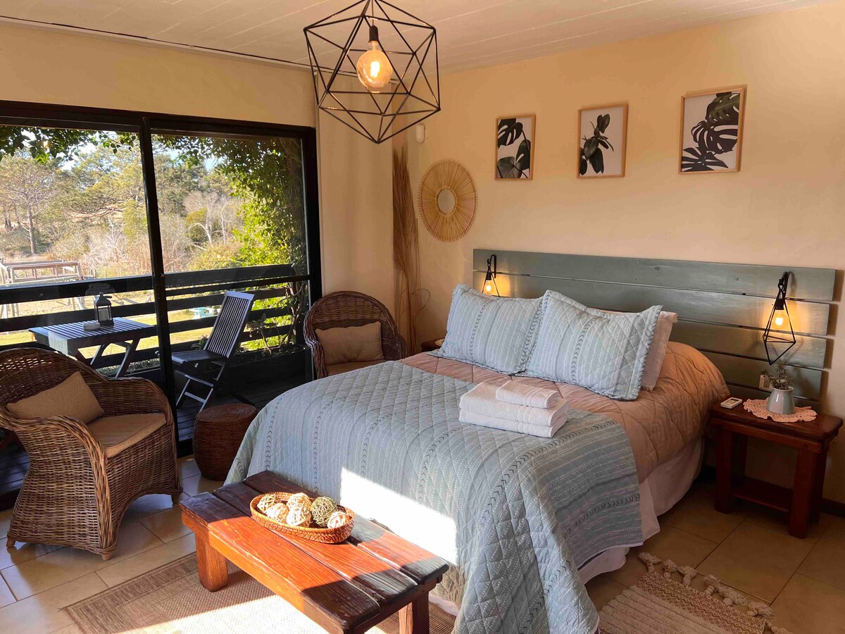 A cozy sleeping area features a spacious bed with soft linens and decorative pillows. A woven rug is positioned in front of the bed. Natural light pours in through large glass doors, framing a view of outdoor seating and greenery.