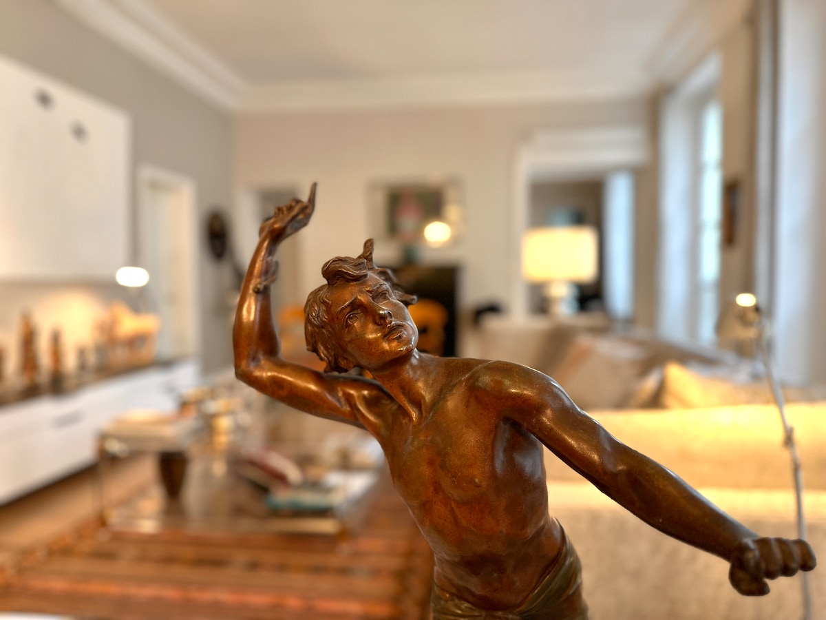 A bronze statue is featured prominently in the foreground, capturing a graceful pose. In the blurred background, a spacious and elegantly furnished living area is visible, with soft natural light filtering through large windows, illuminating a comfortable sofa and decorative elements.